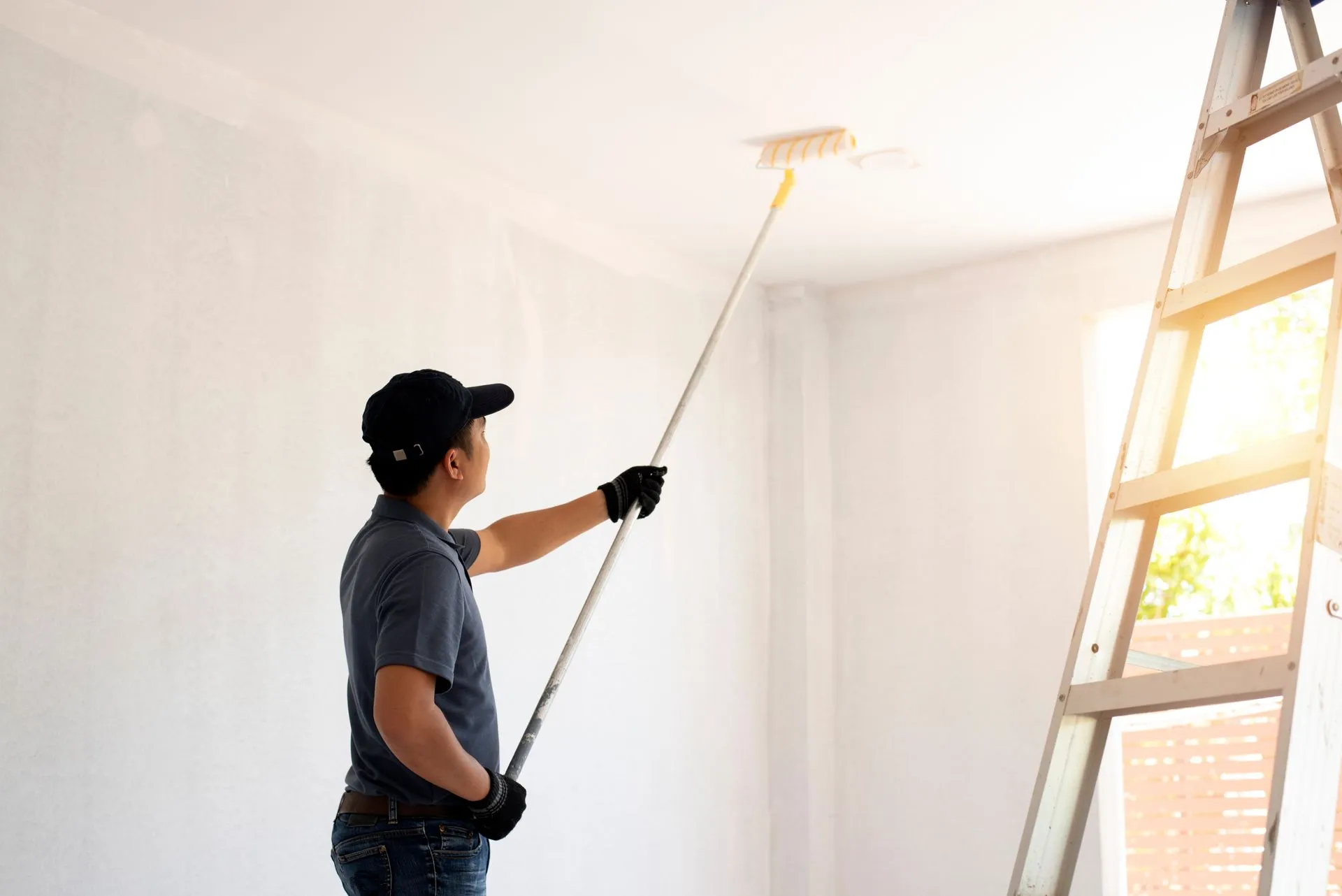 Person paints a white ceiling with a paint roller on an extension pole, near a wooden ladder and window.